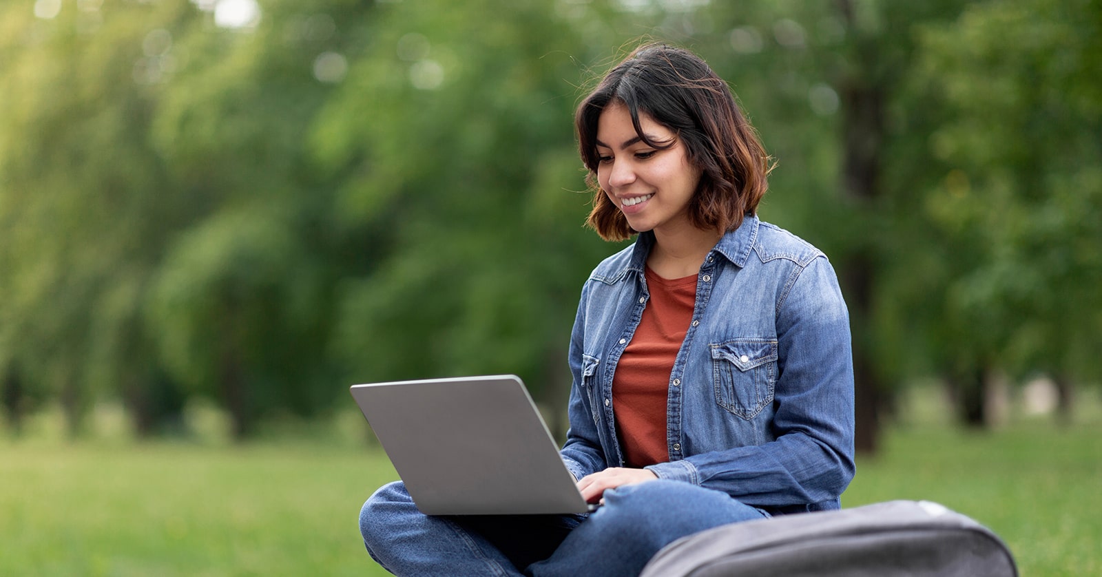 A female college student works outdoors on a laptop, surrounded by trees