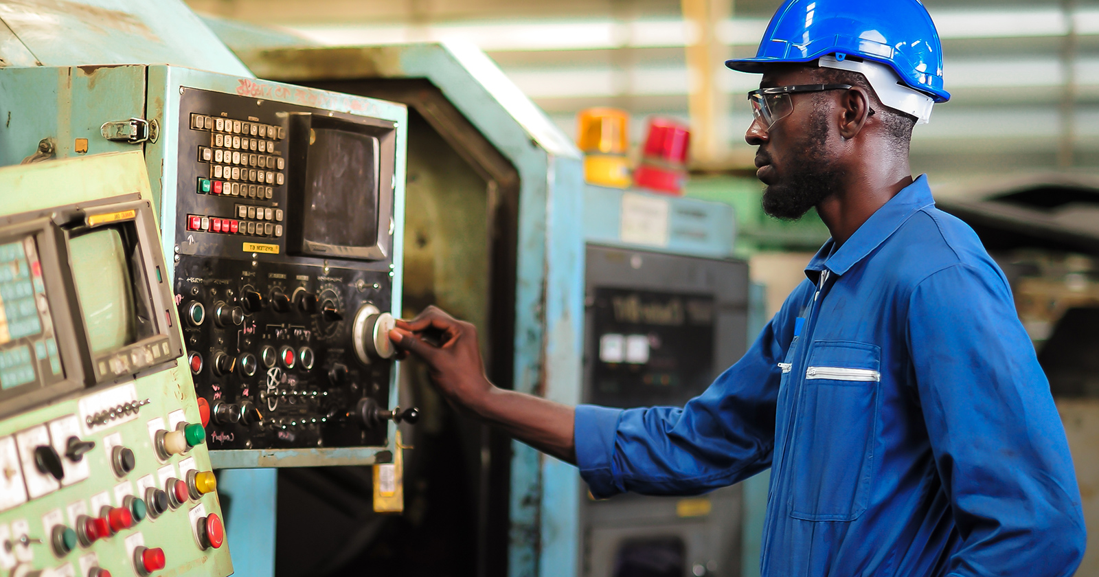 A Black man wearing a blue jumpsuit and hard hat works on a lathe in a factory.