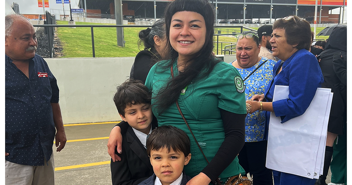 A mother in a work uniform standing proudly behind her two children at an outdoor event.