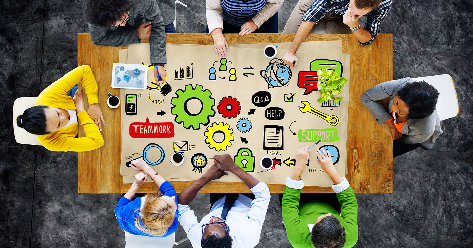 Overhead view of a team of people around a table. The table shows a colorful illustration of gears, charts, and text bubbles.