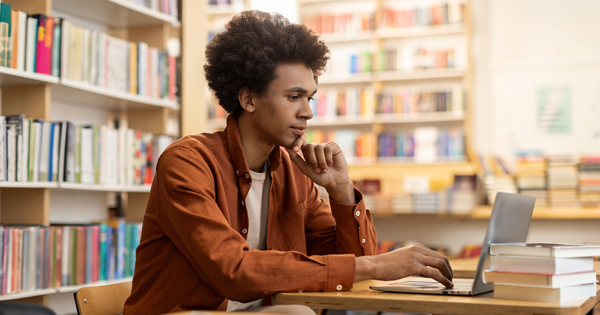 high school student in a library is searching somethig on his laptop