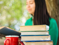 College student shown studying outside, a stack of books and coffee mug in the foreground