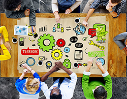 Overhead view of a team of people around a table. The table shows a colorful illustration of gears, charts, and text bubbles.
