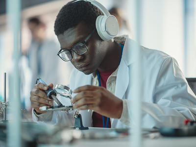 Black male student soldering a circuit board wearing headphones, a lab coat, and goggles