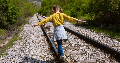A young girl shown from behind balances on the beam a train track in a wooded environment