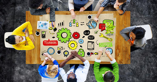 Overhead view of a team of people around a table. The table shows a colorful illustration of gears, charts, and text bubbles.