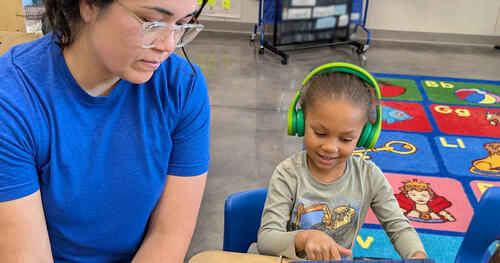 a little girl with a headphone is looking at a tablet with a teacher