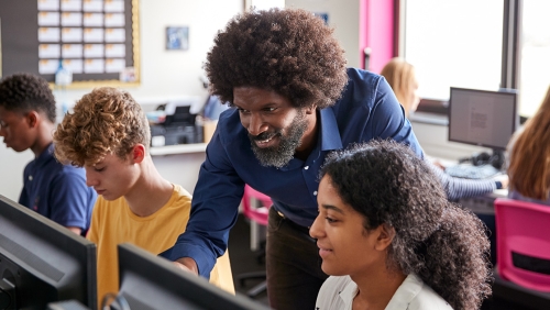 Teacher instructs high school student over her shoulder, looking at a computer together