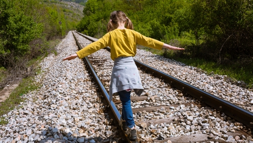A young girl shown from behind balances on the beam a train track in a wooded environment