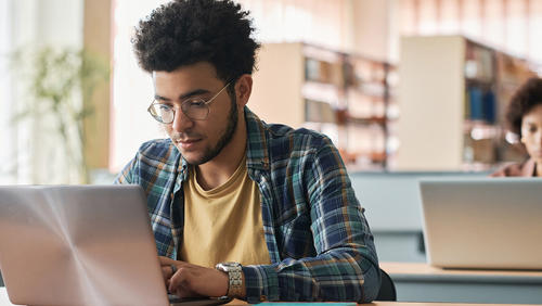Male college student with glasses using a laptop in the library