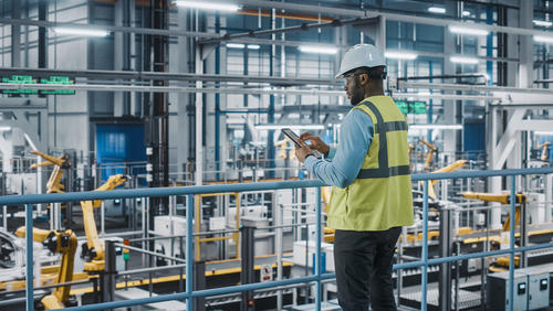 Car factory engineer in a vest and hard hat using a tablet computer and looking at the production line