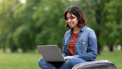 A female college student works outdoors on a laptop, surrounded by trees