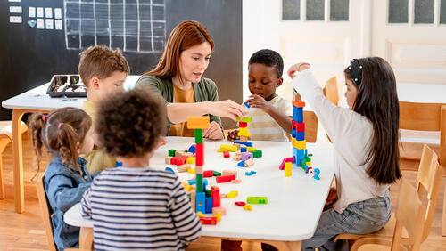 Pre-K instructor assists diverse group of children who are playing with colorful blocks in a classroom
