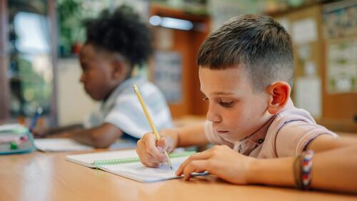 Two first grade boys write in a workbook with pencils 