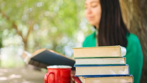College student shown studying outside, a stack of books and coffee mug in the foreground
