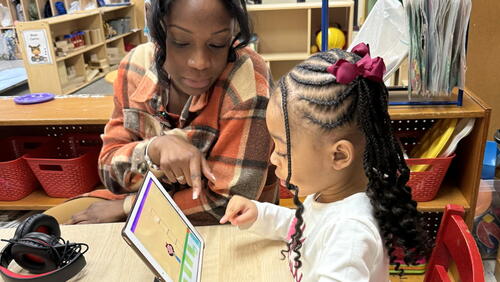 A pre-K child takes an assessment on a tablet in a classroom, supervised by an instructor.