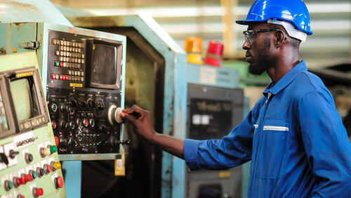 A Black man wearing a blue jumpsuit and hard hat works on a lathe in a factory.