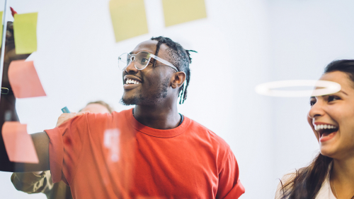 Smiling young Black man writes on sticky notes on a clear board with colleague