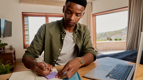 Black male college student takes notes in front of his laptop in a dorm room