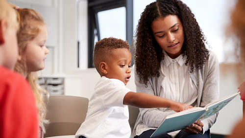 boy showing something in a book holding by teacher with other kids nearby