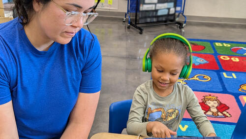 a little girl with a headphone is looking at a tablet with a teacher