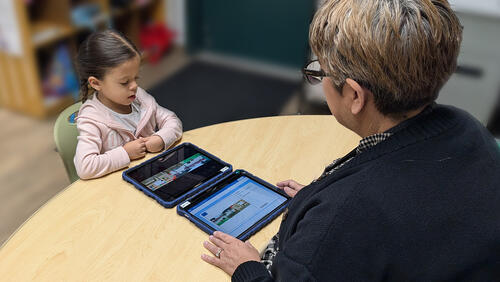A Pre-K student uses a tablet as an educator guides her during an assessment in a classroom