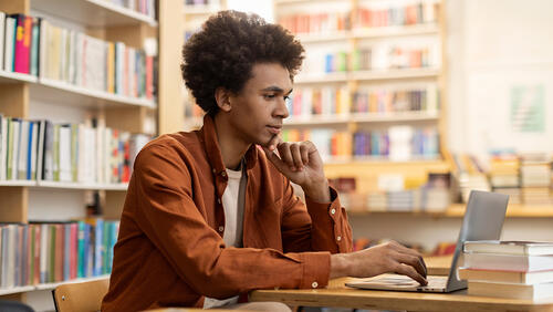 high school student in a library is searching somethig on his laptop