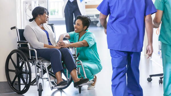 A hospital nurse crouches down to talk with a patient in a wheelchair