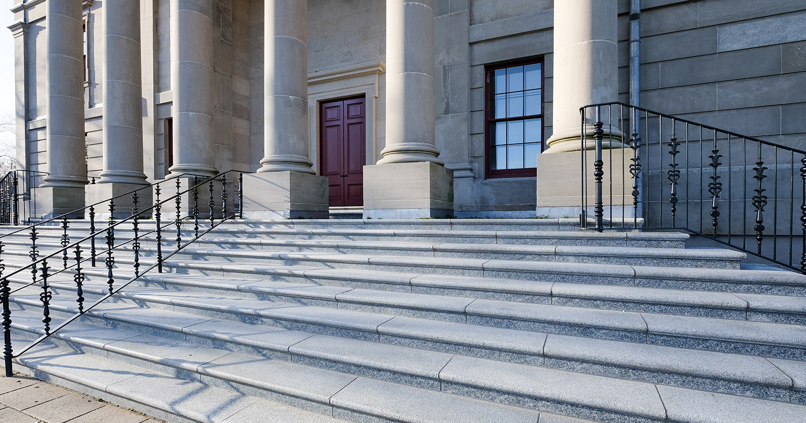 A corner view of a government building with large round pillars, red doors, black metal rails, grey marble steps and brick entrance