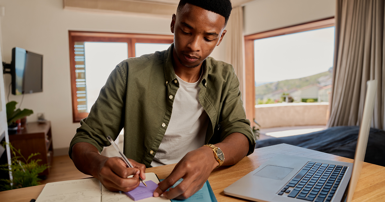 Black male college student takes notes in front of his laptop in a dorm room