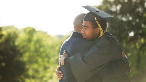 Black college graduate in cap and gown hugging his father