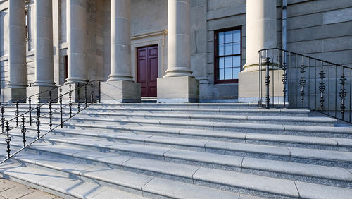 A corner view of a government building with large round pillars, red doors, black metal rails, grey marble steps and brick entrance