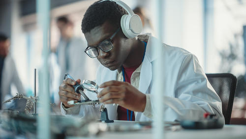 Black male student soldering a circuit board wearing headphones, a lab coat, and goggles