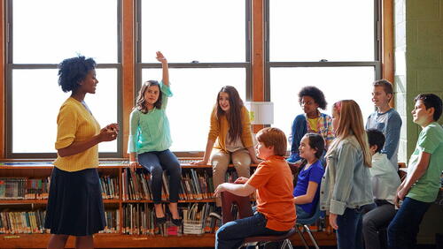 A teacher instructs a diverse group of middle school students in a library.